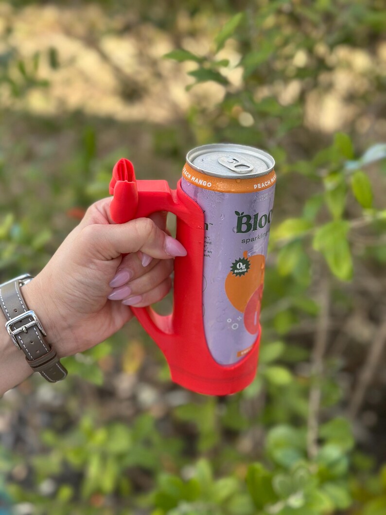 May include: A red 3D-printed can holder with a handle, holding a can of Bloom sparkling water. The can is purple with orange accents and the words "Peach Mango" and "Bloom" are visible. The background is blurred green foliage.