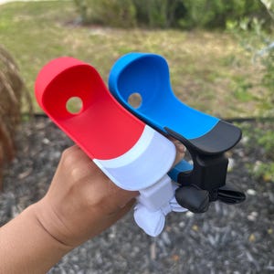 May include: Two plastic boat-shaped accessories, one red and white, the other blue and black. Each has a hole and a clip-like mechanism. The accessories are held up against a blurred outdoor background.