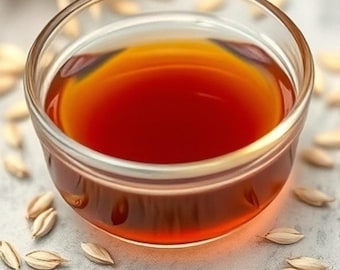 May include: A small glass bowl filled with a dark amber liquid, surrounded by light brown oat groats on a white surface.