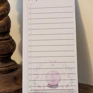 May include: A white notepad with the text "Prophecies & Plans" at the top. The notepad has a series of lines for writing and check boxes. A watercolor illustration of a crystal ball and floral elements adorns the bottom.