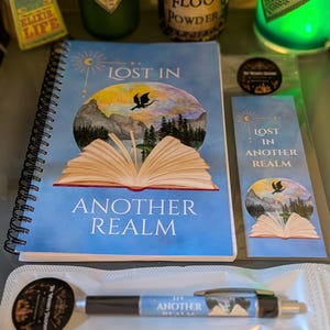 May include: A spiral-bound notebook, a bookmark, and a pen, all featuring the text "LOST IN ANOTHER REALM" and an illustration of an open book with a mountain landscape and a flying dragon silhouette against a circular, colourful sky background.