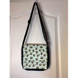 Peut inclure: Un sac bandoulière doté d'un rabat en tissu vert clair avec un motif répétitif de petits personnages extraterrestres verts. Le corps principal du sac et la bandoulière réglable sont en tissu noir uni, contrastant avec le rabat à motifs.