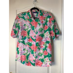 May include: A short-sleeved button-down shirt featuring a vibrant floral pattern in shades of green, pink, and lavender. The shirt has a classic collar, a front button placket, and a label at the neckline that reads HER with a size M indicator.