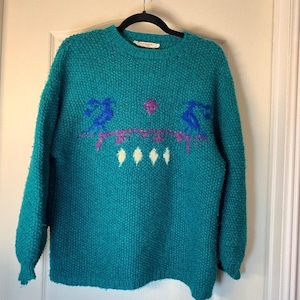 Peut inclure: Pull en tricot de couleur sarcelle avec un col rond et des manches longues. Le pull présente un motif de figures bleues et violettes, un cercle rose et des losanges blancs. Le pull est suspendu à une cintre noire.