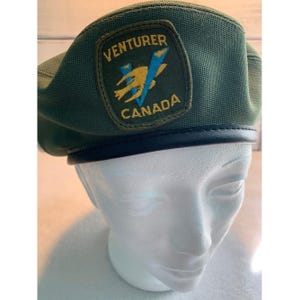 Peut inclure: Un béret vert avec une bande en cuir noir. Le béret présente un écusson avec le texte "VENTURER CANADA" et un emblème jaune et bleu. Le béret est présenté sur une tête de mannequin blanche.