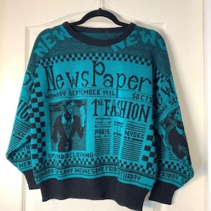May include: Teal and black knit sweater with newspaper-themed graphics. The sweater features the word "NewsPaper" and the text "1st Fashion" in a bold font. Additional text includes "Monday September 14th" and "Splendid Clothing".