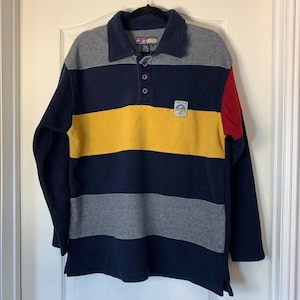 May include: A long-sleeved fleece shirt with a collared neckline and button closure. The shirt features horizontal stripes in navy, grey, and yellow, with a red accent on the right shoulder. A small patch with text is on the chest.