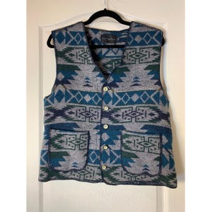 May include: A sleeveless waistcoat featuring a geometric pattern in shades of blue, grey, and dark green. The garment has a V-neckline, five light-coloured buttons down the centre front, and two patch pockets on the lower front section.