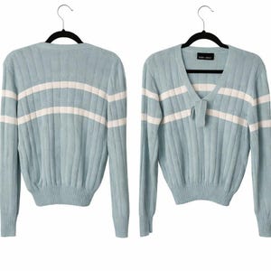 Peut inclure: Un pull en tricot côtelé bleu clair avec des rayures horizontales blanches. Le pull a des manches longues et un col en V avec un petit détail de nœud. Le pull est présenté sur un cintre noir.