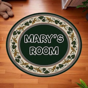 May include: A circular rug featuring the text "MARY'S ROOM" in white against a dark green background. The rug has a decorative border with a beige and green floral pattern. It is placed on a wooden floor.