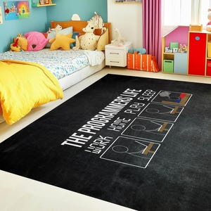 Puede incluir: Alfombra rectangular negra con texto blanco "THE PROGRAMMERS LIFE WORK HOME PLAY SLEEP" y cuatro cuadros ilustrados. La alfombra está en un dormitorio infantil con una cama, juguetes y una decoración colorida.