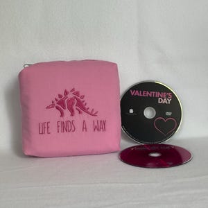 May include: A pink fabric pouch featuring an embroidered dark pink stegosaurus and the text "LIFE FINDS A WAY" in dark pink thread. Next to the pouch are two DVDs, one labeled "VALENTINE'S DAY" with a heart graphic and one translucent red disc.