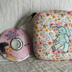 May include: A DVD of "My Big Fat Greek Wedding" with a pink cover and a floral-patterned pillow with a blue bunny applique. The pillow has a white background with pink, orange, and blue flowers.