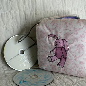 May include: A pink and white floral patterned bag with a purple zip and a stitched pink bunny design. Two CDs are next to the bag, one with the text "Christina Aguilera" and the other with a blue swirl design.