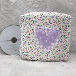 May include: A square fabric pouch featuring a white background with a small floral print in shades of purple, pink, and green. The center is decorated with a lavender heart appliqué that has white polka dots and a light purple gingham border.
