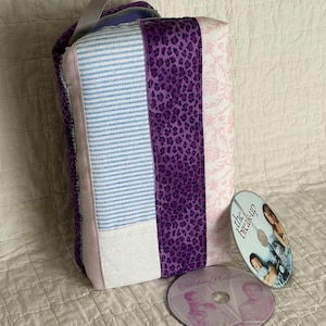 以下が含まれることがあります： グレーの持ち手が付いた長方形のパッチワークバッグ。青と白のストライプ、紫のヒョウ柄、ピンクの花柄のパネルがあります。「The Break-Up」と「Wedding Planner」の2枚のDVDがバッグの横にあります。