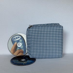 Puede incluir: Un estuche cuadrado de tela con estampado de cuadros vichy en azul y blanco con cierre de cremallera, colocado junto a dos discos. Un disco muestra un gráfico de mando a distancia con el texto CLICK y el otro el texto ENDER'S GAME.