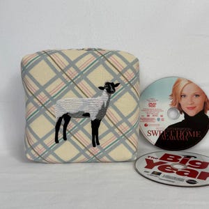 May include: A square fabric cushion featuring a cream-colored background with a light blue, pink, and grey plaid pattern and an embroidered black and white sheep in the center, positioned next to two DVDs titled Sweet Home Alabama and The Big Year.