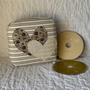May include: A decorative square pillow with a striped pattern and three heart appliqués. Two hearts feature a floral design, and one is a solid cream color. Two DVDs are next to the pillow.
