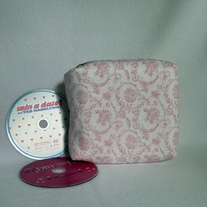 Könnte beinhalten: Ein weicher, rechteckiger Stoffbeutel mit weißem Hintergrund und einem sich wiederholenden rosa Blumenmuster. Neben dem Beutel liegen zwei DVDs: eine mit dem Titel Win a Date with Tad Hamilton mit blauen Sternen und Herzen und eine mit 27 Dresses.
