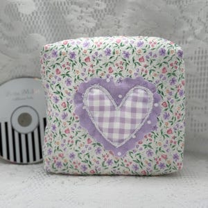 May include: A square fabric cushion featuring a white background with a small-scale floral print in shades of pink, purple, and green. Centered on the front is a lavender heart appliqué with a white gingham pattern and a scalloped edge.