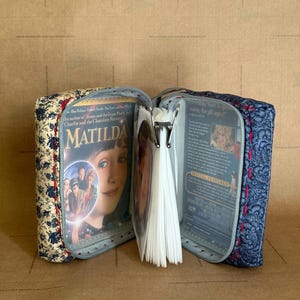 Puede incluir: Estuche de DVD abierto con un estampado floral. El estuche está abierto, revelando la portada del DVD de la película "Matilda". El estuche es azul y blanco con una cremallera y costuras rojas.