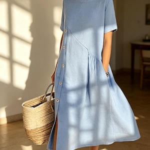 May include: A light blue linen dress with short sleeves and a button-down front. The dress features a side slit and a gathered detail at the waist. A woven straw tote bag is held in the hand.