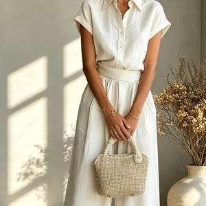 May include: A cream-colored linen outfit consisting of a short-sleeved button-down shirt and wide-leg pants. A woven handbag with a circular clasp is held in front. The outfit is accessorized with gold bracelets.