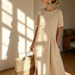 May include: A beige linen dress with short sleeves and a button-up front. The dress has a side slit and is paired with a woven tote bag. The dress is a neutral color and appears to be lightweight.