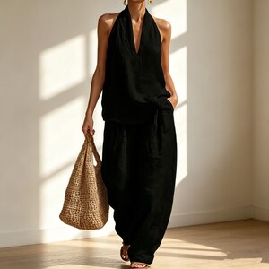 May include: A black halter top and wide-leg pants set. The top has a deep V-neck. A woven tote bag is carried in the left hand. The outfit is paired with sandals. The outfit is made of a lightweight fabric.