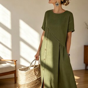 May include: Olive green linen dress with a button-down front and short sleeves. The dress features a side slit and is paired with a woven tote bag. The dress is approximately knee-length.