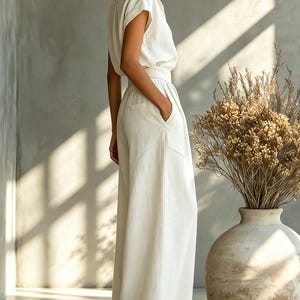 May include: A cream-colored linen jumpsuit with a collared, short-sleeved top and wide-leg pants. The outfit is belted at the waist and has pockets. The model is wearing sunglasses and standing near a large vase with dried plants.