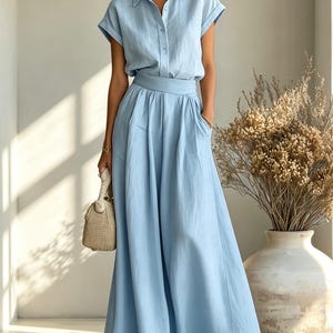 May include: A light blue two-piece outfit featuring a button-down shirt with short sleeves and a matching long skirt. The skirt has a flowing design with pockets. A small woven handbag and sandals complete the look. The outfit is made of a lightweight fabric.