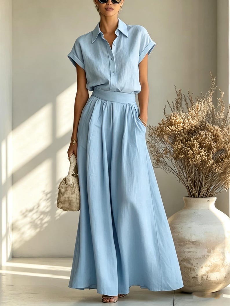 May include: A light blue two-piece outfit featuring a button-down shirt with short sleeves and a matching long skirt. The skirt has a flowing design with pockets. A small woven handbag and sandals complete the look. The outfit is made of a lightweight fabric.