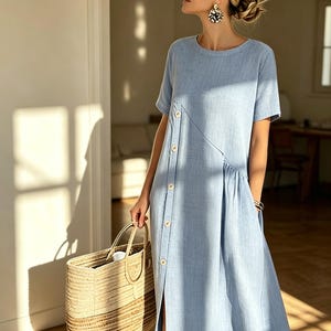 May include: A light blue midi dress with short sleeves and a button-up front. The dress features a side slit and a round neckline. A woven straw tote bag is held in the hand. The dress is made of a lightweight fabric.