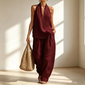 May include: A burgundy halter top and wide-leg pants set. The top has a deep V-neck, and the pants have a relaxed fit. A woven tote bag is held in the hand. The outfit is paired with sandals. The clothing is made of a lightweight fabric.
