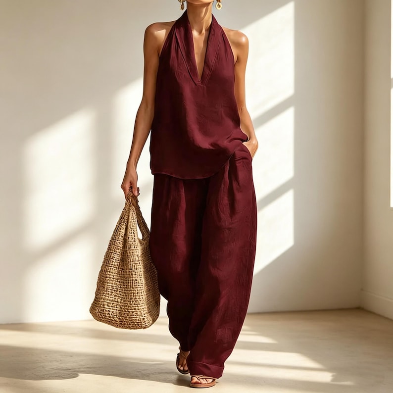 May include: A burgundy halter top and wide-leg pants set. The top has a deep V-neck, and the pants have a relaxed fit. A woven tote bag is held in the hand. The outfit is paired with sandals. The clothing is made of a lightweight fabric.
