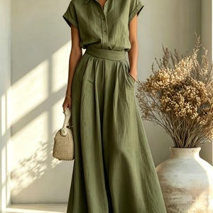 May include: Olive green two-piece outfit featuring a short-sleeved button-down shirt and a flowing maxi skirt. The outfit includes a matching belt and pockets. A woven handbag and sandals complete the look. A large vase with dried plants is in the background.