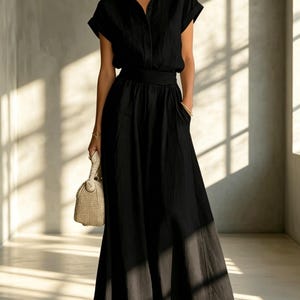 May include: A black dress featuring a collared, button-down top with short sleeves and a flowing maxi skirt. The dress has a defined waist and pockets. A beige handbag is held in one hand.