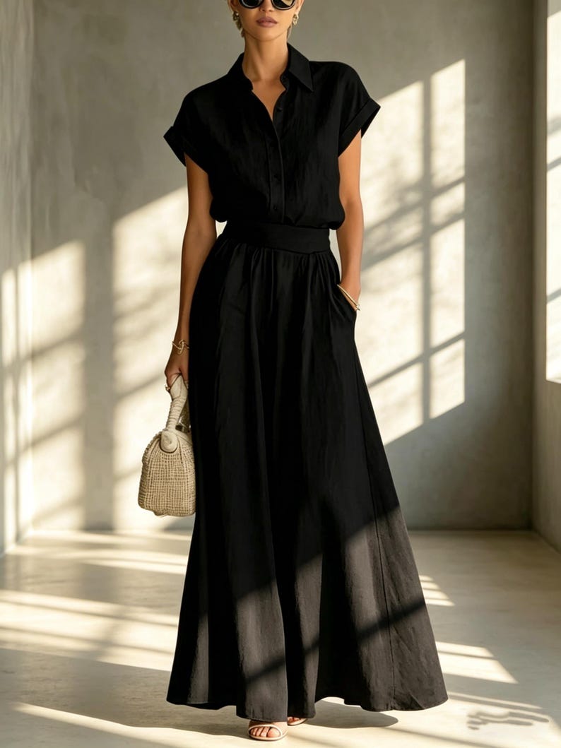 May include: A black dress featuring a collared, button-down top with short sleeves and a flowing maxi skirt. The dress has a defined waist and pockets. A beige handbag is held in one hand.