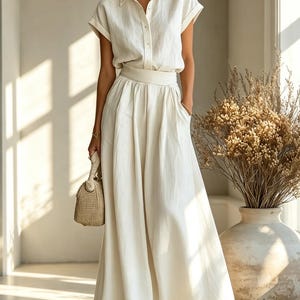 May include: A cream-colored linen two-piece set consisting of a short-sleeved button-down shirt and a flowing maxi skirt. The outfit is paired with a woven handbag and sandals. The ensemble is suitable for warm weather and casual occasions.