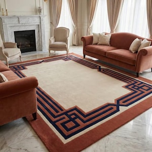 May include: A large, rectangular rug with a cream-colored center and a geometric border in shades of rust, navy blue, and cream. The rug is placed in a living room with a fireplace, sofas, and chairs.