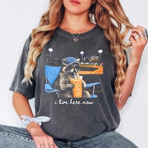 May include: Dark gray t-shirt featuring a raccoon wearing sunglasses and a baseball cap, sitting in a blue chair, and drinking an iced coffee. The graphic includes a baseball field background and the text "i live here now."