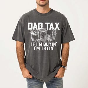 May include: Dark grey t-shirt with the text "DAD TAX" above an illustration of fast food items. Below the text is "IF I'M BUYIN' I'M TRYIN'". The shirt has a vintage, worn look.