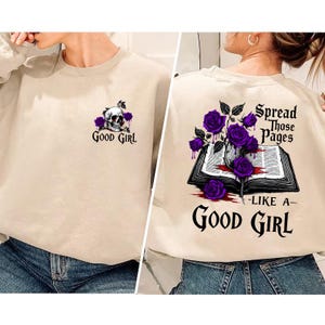 May include: Beige sweatshirt with a skull and purple roses graphic, and the text "Good Girl" on the front. The back features a book with purple roses, the text "Spread Those Pages - Like a - Good Girl".