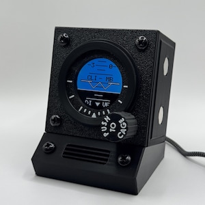 Puede incluir: Un instrumento de aviación negro de forma rectangular con un indicador circular que muestra una línea de horizonte azul y negra. El indicador muestra el texto CLIMB y DIVE. Un mando negro con la etiqueta PUSH TO CAGE está en el panel frontal.