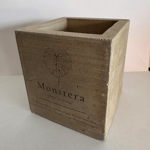 May include: A square, light brown concrete planter with the word "Monstera" and a leaf illustration. The planter has a textured surface and is designed for indoor or outdoor use. The planter is 15 cm tall and 13 cm wide.