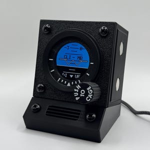 Könnte beinhalten: Ein schwarzes, rechteckiges Instrument im Luftfahrt-Design mit strukturierter Oberfläche. Die kreisförmige Anzeige zeigt eine blaue und schwarze Horizontlinie mit dem Text CLIMB und DI-UF. Ein schwarzer Knopf mit der Aufschrift PUSH TO CAGE ist vorne.