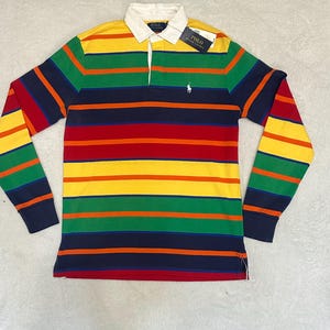 Puede incluir: Una camiseta de rugby de manga larga con rayas horizontales amarillas, verdes, naranjas, rojas y azul marino. La camiseta tiene un cuello blanco y un pequeño logotipo bordado de un jugador de polo. Una etiqueta está adherida a la camiseta.
