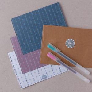 Puede incluir: Un surtido de artículos de papelería, que incluyen hojas de papel estampadas en azul, morado y blanco, un sobre marrón con una pegatina circular y dos bolígrafos de gel con tinta turquesa y rosa. El papel presenta un diseño floral.